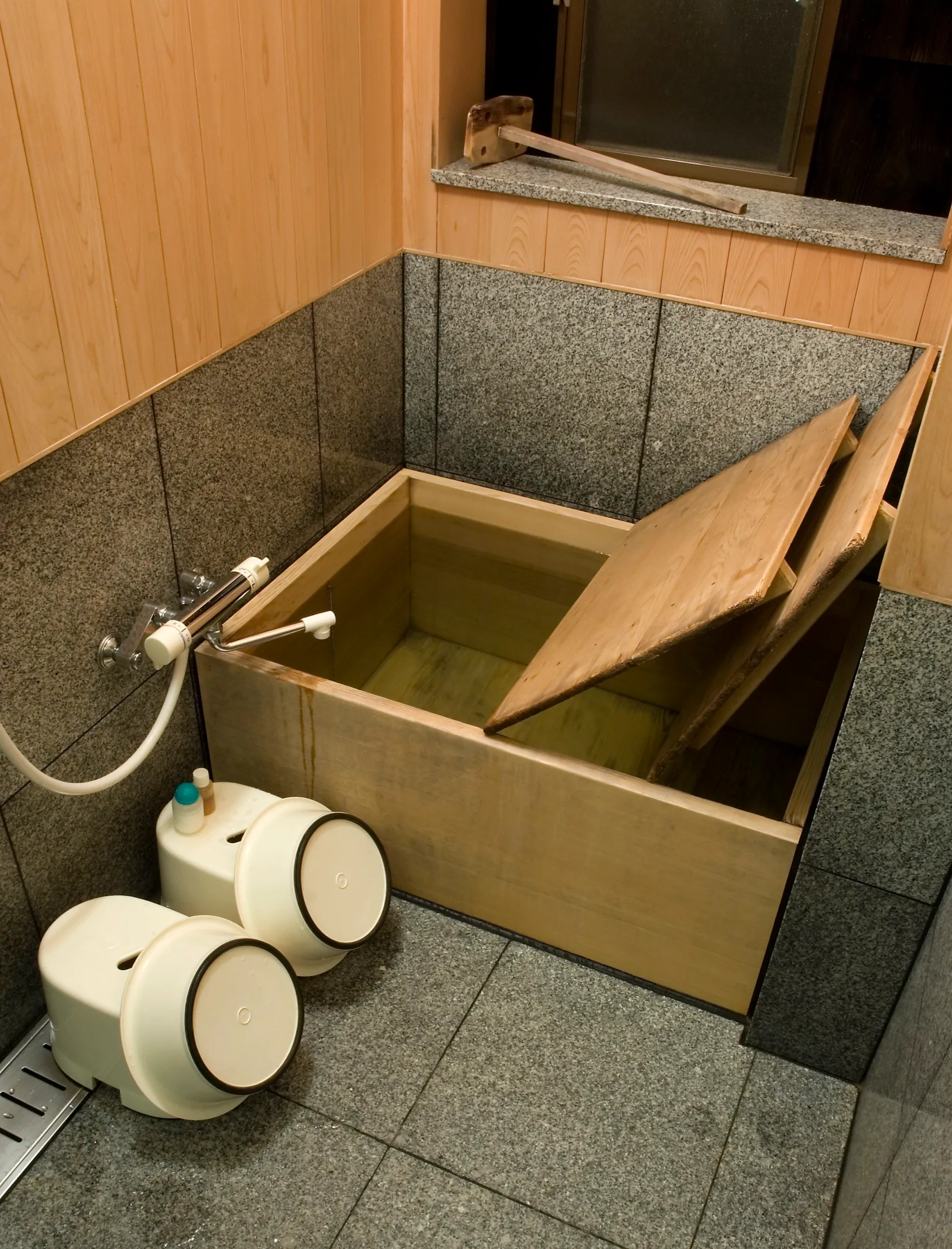 Ofuro de madera clara en un baño japonés de piedra, con la tapa de madera entreabierta